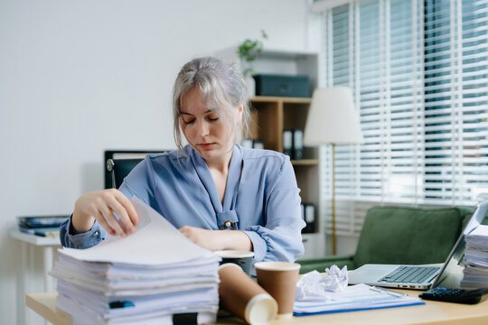 Focused businesswoman sorting documents at a cluttered desk with reports, coffee cups, and files. Perfect for accounting, finance, tax season, and deadline stress concepts. - Powered by Adobe
