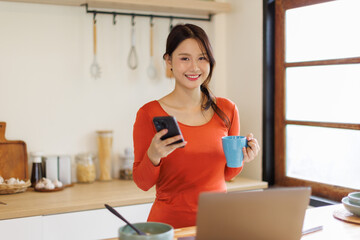 Happy asian woman drinking tea while using smart phone in the kitchen. Copy space.