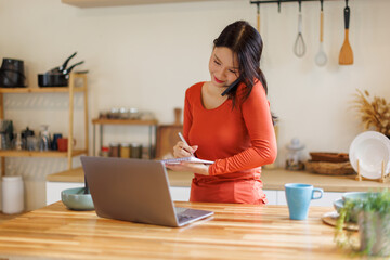 Asian woman checking laptop talking on phone in a kitchen at home
