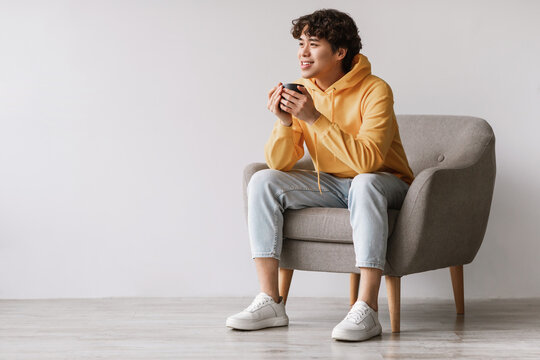 Cheerful Asian man sits comfortably in a gray armchair, sipping coffee. He enjoys his free time in a bright studio with white walls, showcasing a relaxed atmosphere.
