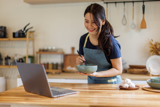 Woman, cooking in kitchen with laptop for live streaming, online recipe and meal prep by stove. Nutrition influencer, food blogger or vegan chef with social media tutorial in apartment
