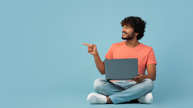 A handsome Indian man in a casual outfit sits on the floor, using a modern laptop. He is typing and pointing at a blue background, portraying focus and creativity while working online.