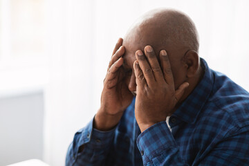 Mature African man is sitting indoors, visibly distressed as he touches his head. The side view captures his struggle with a painful headache, reflecting unhappiness and health issues.