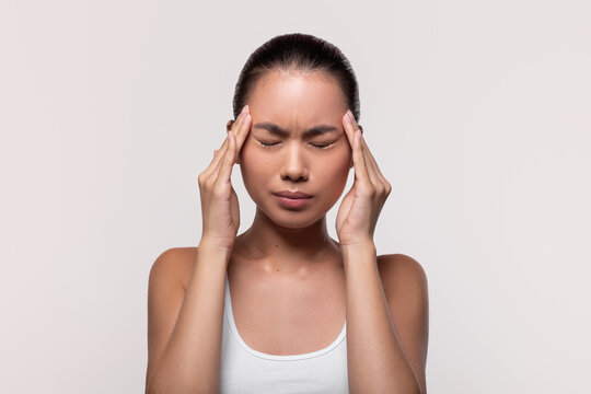 Unhappy Chinese woman in her twenties is massaging her temples with closed eyes, experiencing discomfort from a migraine. The grey background provides a calm setting for her struggle.
