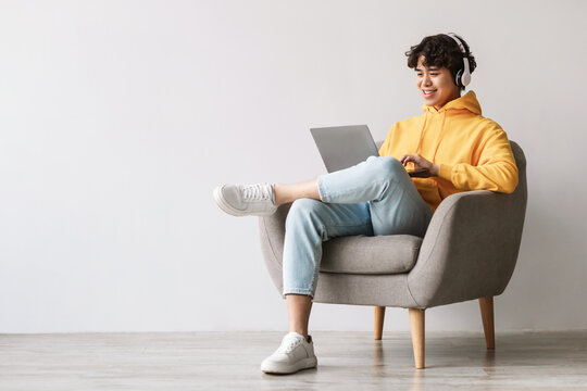 Happy young Asian man sits in a gray armchair, wearing headphones and using a laptop. He communicates online while enjoying a relaxed atmosphere against a white wall.