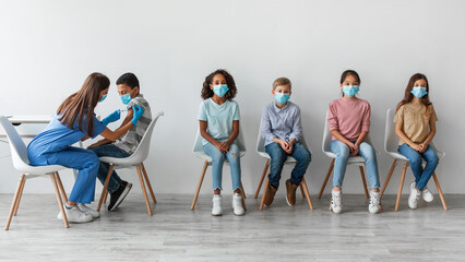 Children sit patiently as a female nurse administers Covid-19 vaccinations. The diverse group includes multiracial boys and girls in a neutral background. Their health is being prioritized.