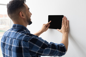 A joyful millennial man is using a digital tablet on a wall to control his home automation system. He is adjusting settings while standing indoors, enjoying innovative technology.