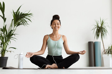 A young woman sits in a lotus position on a mat in a bright yoga studio. She is smiling and enjoying music through earphones while meditating. Plants are visible in her surroundings.
