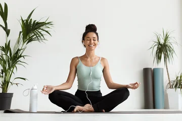 Fotobehang Lotusbloem A young woman sits in a lotus position on a mat in a bright yoga studio. She is smiling and enjoying music through earphones while meditating. Plants are visible in her surroundings.  © Prostock-studio