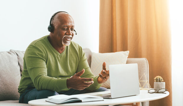 A senior man is sitting on a couch in a cozy living room, smiling and talking on a laptop. He wears headphones and gestures with his hands, engaged in a lively discussion.