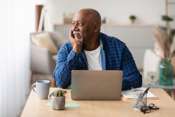 Mature African American businessman is sitting at a desk with a laptop in a modern office. He looks thoughtfully aside while engaged in work, creating a serene atmosphere.