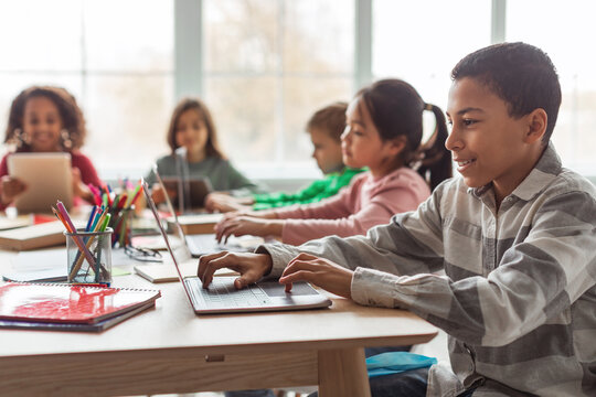 In a modern classroom, an African American schoolboy uses a laptop for online learning while sitting with his multicultural classmates. They are focused on their educational tasks.