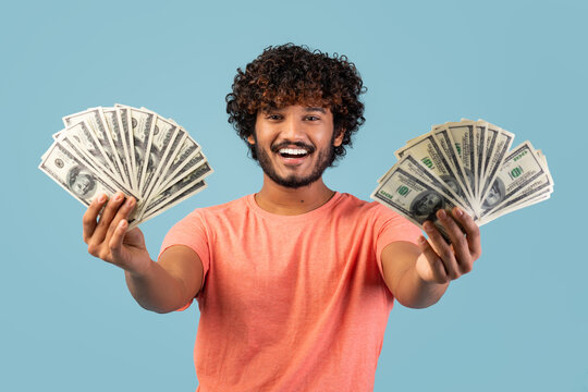 A joyful Indian man in a pink t-shirt holds cash in both hands while smiling at the camera. He showcases his excitement against a bright blue studio backdrop, embodying happiness and success.
