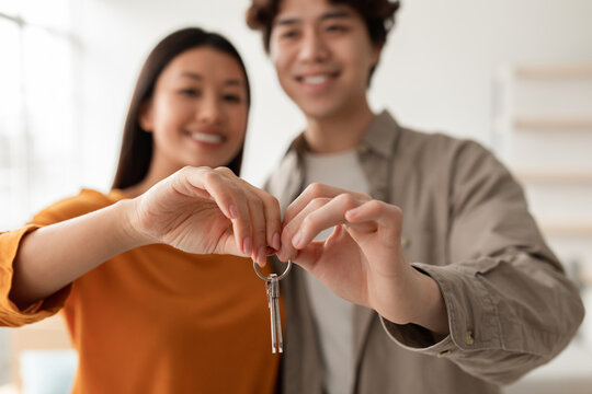 Joyful millennial Asian couple shares a house key on moving day. They embrace their new home together while showing love and excitement in their cozy apartment space.