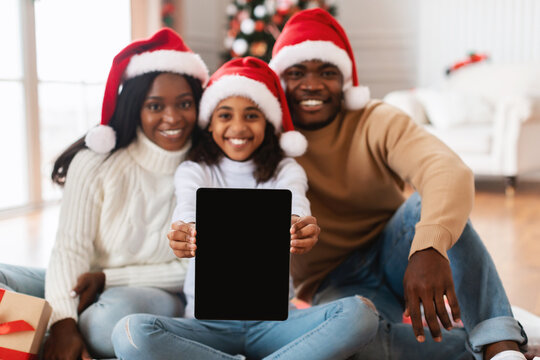 Joyful black family in red Santa hats is sitting on the floor, showing a tablet with an empty screen. They celebrate Christmas together, surrounded by holiday decorations and gifts. - Powered by Adobe