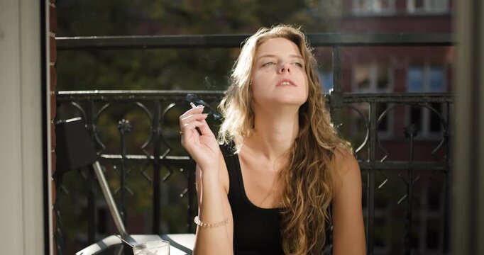 Girl relaxing on balcony enjoying cigarette while basking in the sun outside an apartment in the city during a pleasant afternoon