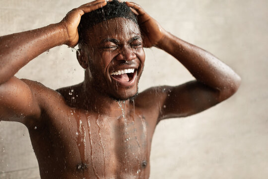 A black man stands under a shower in a modern bathroom, eyes closed and smiling as the water cascades down. He is focused on his refreshing shower experience, enjoying his grooming routine.