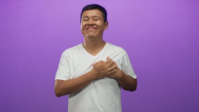 Man smiling with eyes closed holding hands to chest in studio against purple backdrop with soft smile; gratitude.