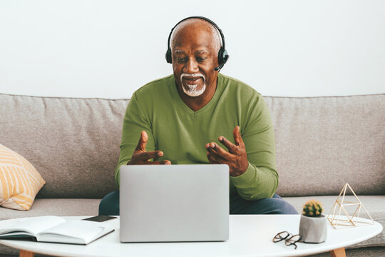 A senior man wearing a headset actively participates in an online discussion from his comfortable couch. He gestures expressively while focusing on the laptop screen in his modern living space.