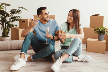 A happy couple enjoys each other's company and drinks, sitting on the floor in their new home. Surrounding them are moving boxes and indoor plants, creating a cozy atmosphere of celebration.