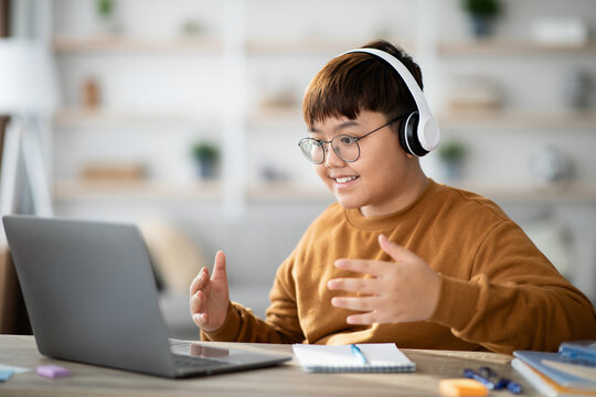 A chubby Asian boy is actively attending an online lesson from his home. He uses a laptop and wears a wireless headset, showing excitement while working on his educational project.