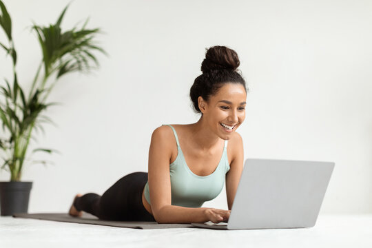 A young woman in athletic clothing lies comfortably on a fitness mat, focused on her laptop. She smiles happily while browsing online, enjoying her time at home with a green plant nearby.