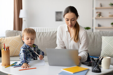 A woman assists her young son as he draws on a table filled with art supplies. She is focused on...