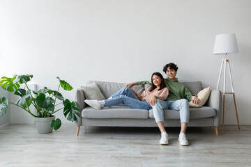 Obraz premium In a cozy living room, a young Asian couple cuddles on a light gray couch, sharing warm moments. They relax amidst a bright and spacious atmosphere, surrounded by plants.
