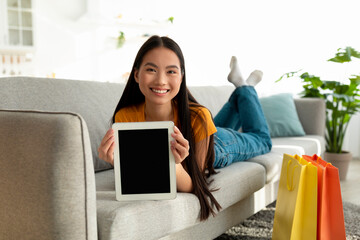 A cheerful Asian woman lies on a sofa, holding a digital tablet with a blank screen. Surrounded by shopping bags, she smiles as she engages in online shopping from her comfortable home setting.