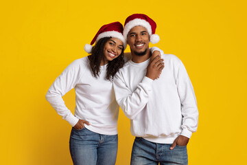 Two friends are smiling and posing together, wearing matching white sweaters and red Santa hats. The bright yellow background adds a cheerful holiday vibe to the scene.