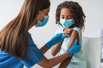 A medical worker is giving a Covid-19 vaccination to a young African girl in a clinic indoors. Both are wearing face masks for safety during the antiviral immunization.