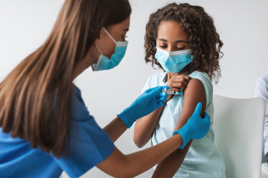 A medical worker is giving a Covid-19 vaccination to a young African girl in a clinic indoors. Both are wearing face masks for safety during the antiviral immunization. - Powered by Adobe
