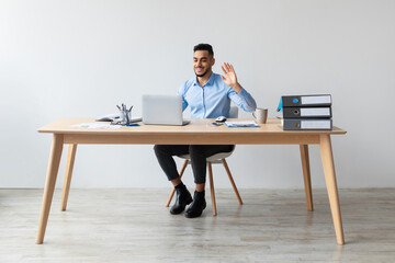 In a bright office, a young Arab businessman sits at a wooden desk using a laptop for a video call. He is smiling and waving his hand, engaging in online communication.