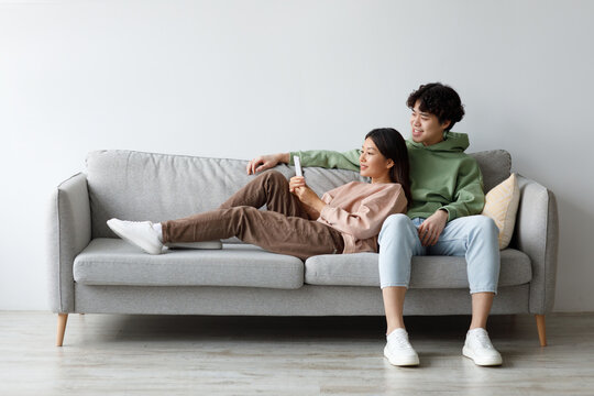 Young Asian couple enjoys quality time on a comfortable couch at home, happily browsing a smartphone and exploring the internet. They share a relaxing moment, engaged with each other.