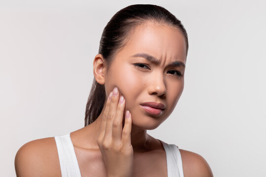Unhappy asian millennial lady is experiencing severe wisdom tooth pain. She touches her cheek with a worried expression. The background is white and provides empty space for text.