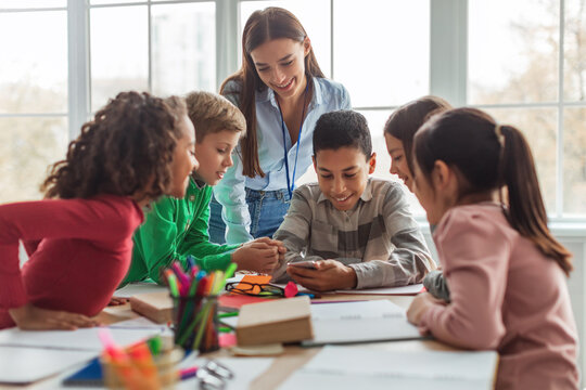 In a modern classroom, diverse school kids gather around their teacher, excitedly using a smartphone for learning. They explore educational applications while seated at a desk.