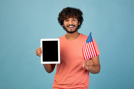 A young eastern man wearing a casual outfit holds an American flag in one hand and a digital tablet with a blank screen in the other. He stands against a blue studio background, smiling cheerfully.