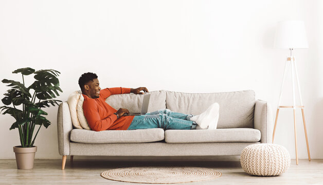 A young man sits comfortably on a light-colored couch in a stylish living room. He works on his laptop while smiling, surrounded by plants and natural light.