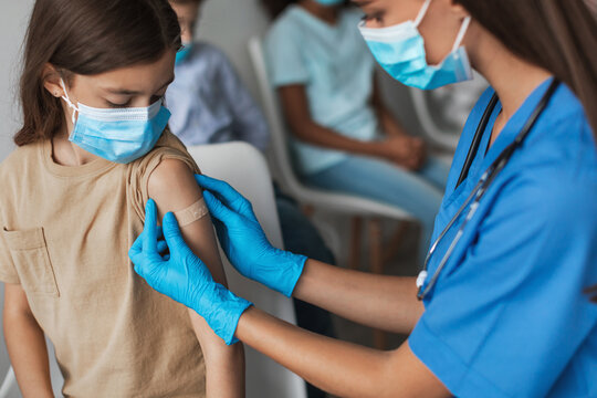 Preteen girl sits in a clinic as a nurse administers a covid-19 vaccine. The nurse carefully applies a medical plaster to the girl's arm following the injection. Other children wait nearby.