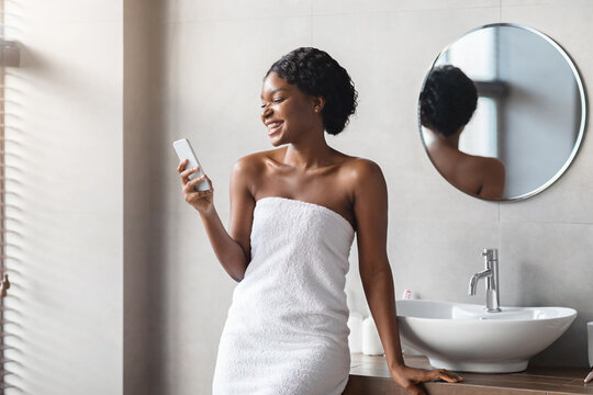 Young black woman wrapped in a towel smiles while using her smartphone in a stylish bathroom. She enjoys a moment of connection during her morning beauty routine, standing by the sink and mirror.