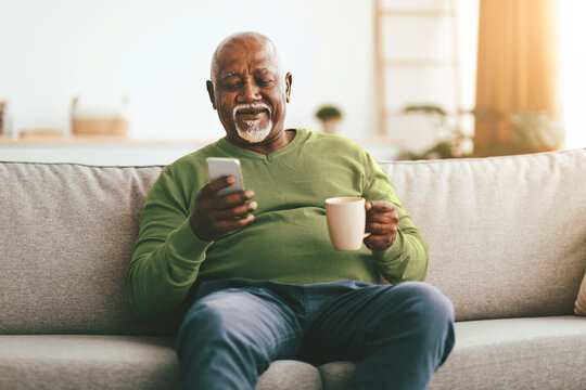 An elderly man sits comfortably on a soft couch, holding a cup of coffee in one hand while looking at his smartphone. Sunlight fills the room, creating a cozy atmosphere.