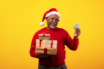 Cheerful senior black man wearing a Santa hat stands in front of an orange background, holding a credit card and a pile of Christmas gifts, enjoying festive holiday shopping.