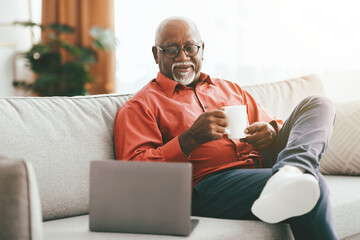 A senior man sits comfortably on a sofa, sipping coffee from a white mug. He is engaged with a laptop in front of him, showcasing a moment of relaxation at home.