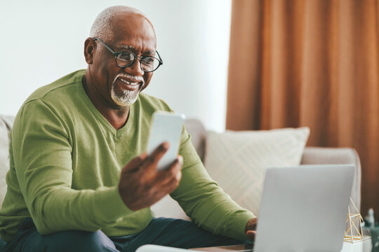 A smiling elderly man sits comfortably on a couch, holding his smartphone and looking at it while a laptop rests in front of him. Natural light fills the modern living room.