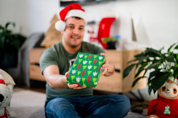 A man wearing a Santa hat happily shows a wrapped present in a festive setting at home.