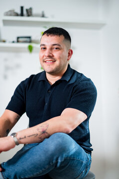A cheerful gay man in his thirties sits comfortably, showing off his tattoos and style