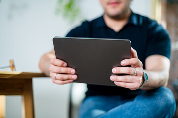 A man in his 30s sits relaxed at home, holding a tablet and engaging with digital content