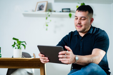 A man in his 30s is sitting in a chair, engaging with a tablet in a cosy home setting