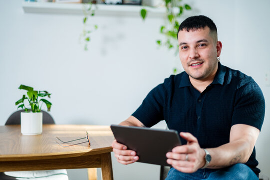 Man in his thirties smiles while using a tablet, seated at a wooden table in a bright room.