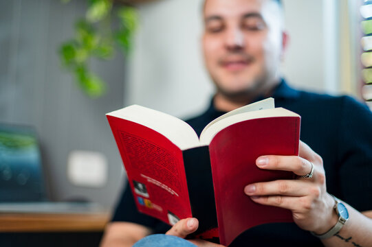 A  man in his 30s reads a novel while sitting comfortably in a well-lit home office
